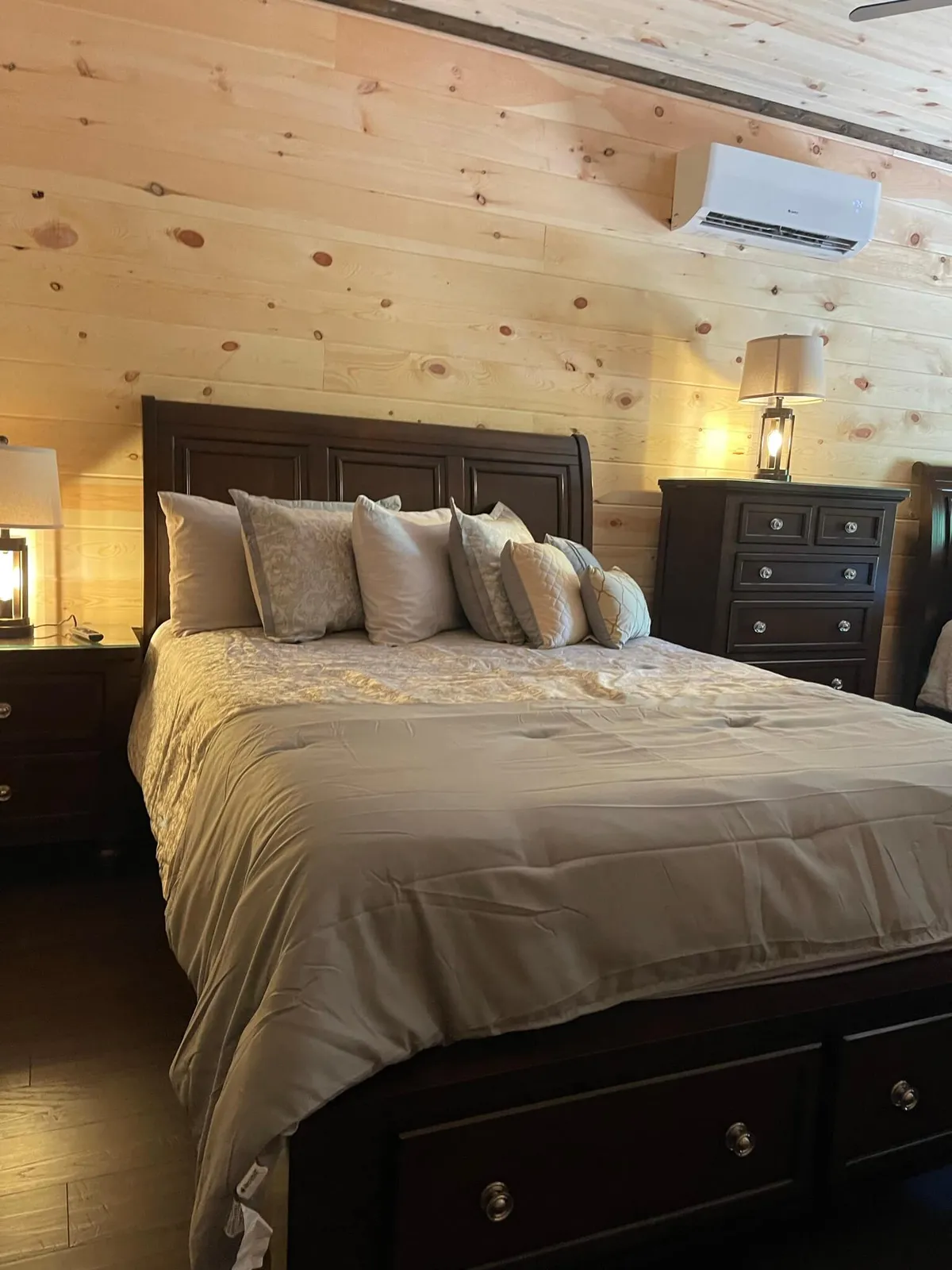 A tranquil bedroom featuring a plush bed with decorative pillows, flanked by nightstands and softly glowing lamps. The wooden wall and gentle morning light create a cozy, restful atmosphere ideal for unwinding.