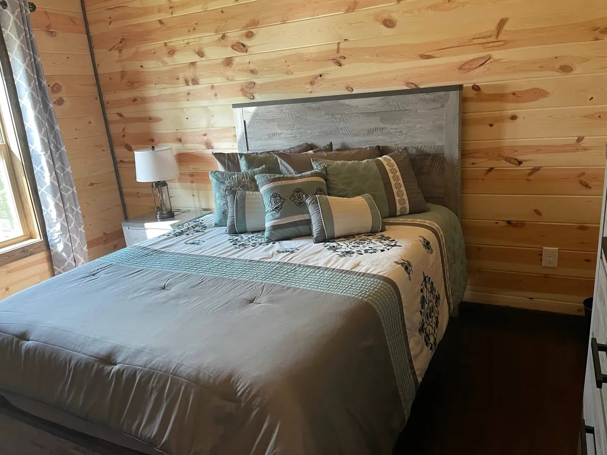 A cozy bedroom with a queen bed, decorative pillows, rich wood paneling, patterned curtains, and a bedside lamp casting a soft glow. The ambiance is peaceful and inviting.
