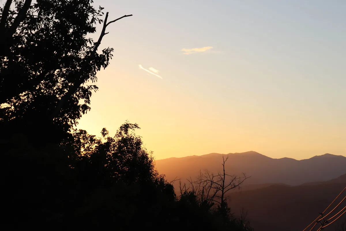 The hero image showcases a stunning mountain range bathed in golden-hour light, with silhouetted trees framing the view. The expansive sky is tinged with vibrant sunset colors, evoking a sense of tranquility and awe. The scene suggests a serene atmosphere perfect for relaxation or quiet reflection, inviting viewers to imagine themselves unwinding in this peaceful mountain retreat.