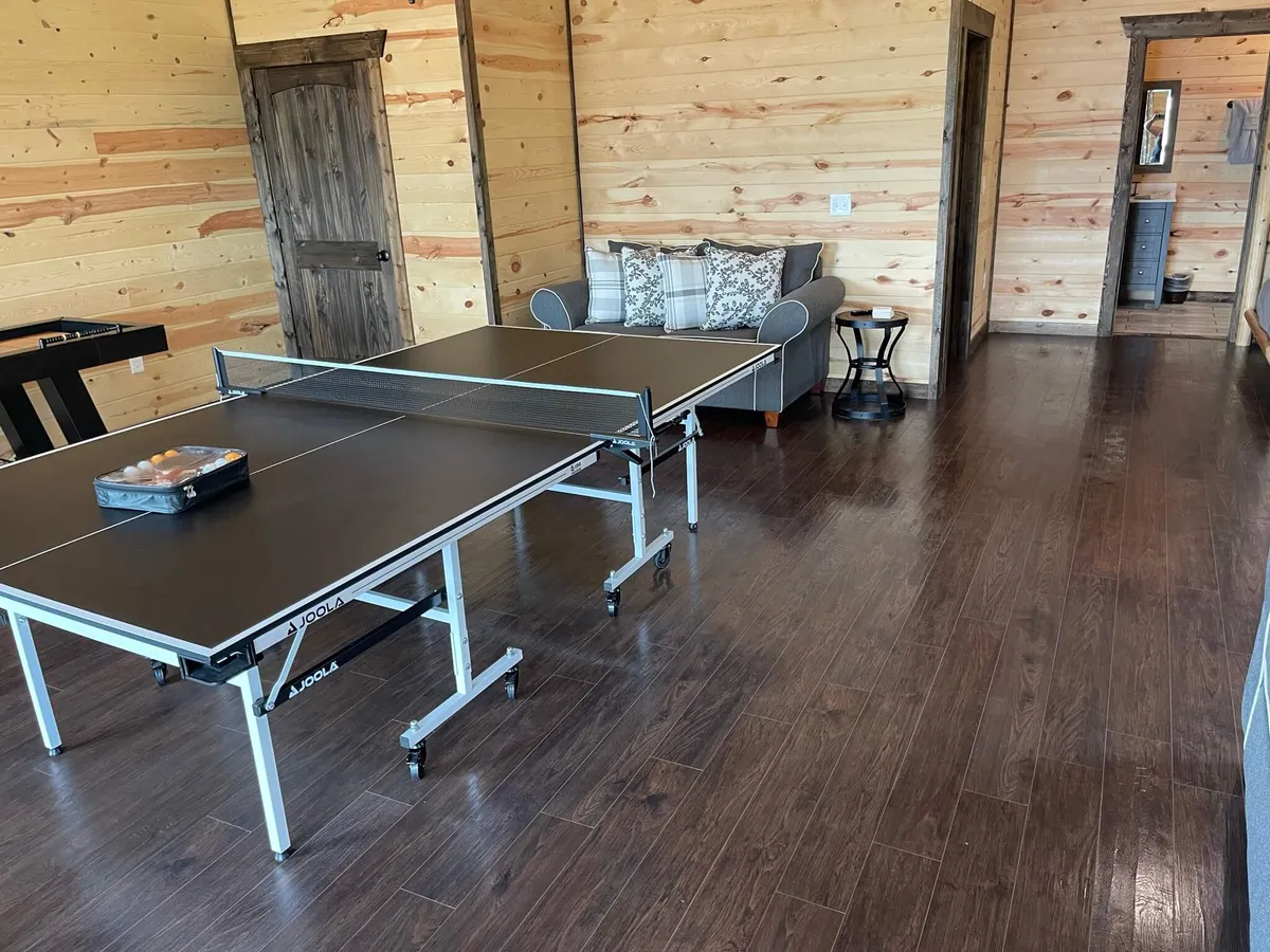 This image features two ping pong tables at the center of a spacious, wood-accented game area. Comfortable seating is arranged nearby, and friends are seen playing and chatting, creating a lively, playful atmosphere. The wood paneling adds warmth and a rustic cabin vibe.