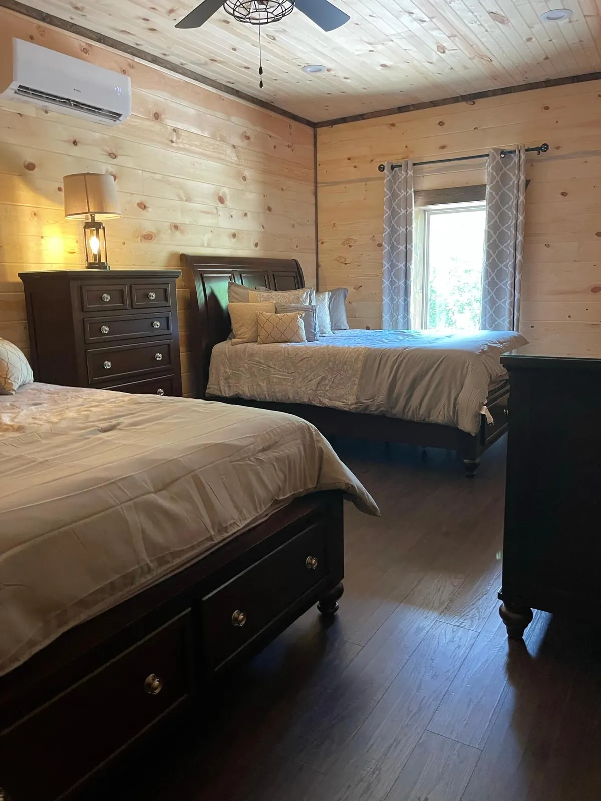A welcoming bedroom with two queen beds, a shared dresser, and a soft lamp. The window lets in natural light, highlighting the inviting, shared space perfect for kids, teens, or friends.