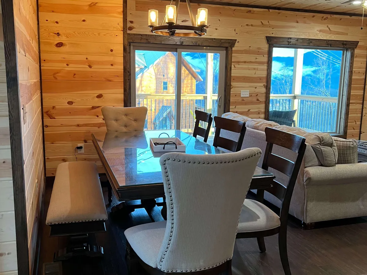 This image presents an inviting open-concept living and dining space featuring a comfortable sofa, a large wooden dining table, an elegant chandelier, and expansive windows that bathe the area in natural light. The windows offer a glimpse of the outdoors, suggesting stunning mountain views beyond.