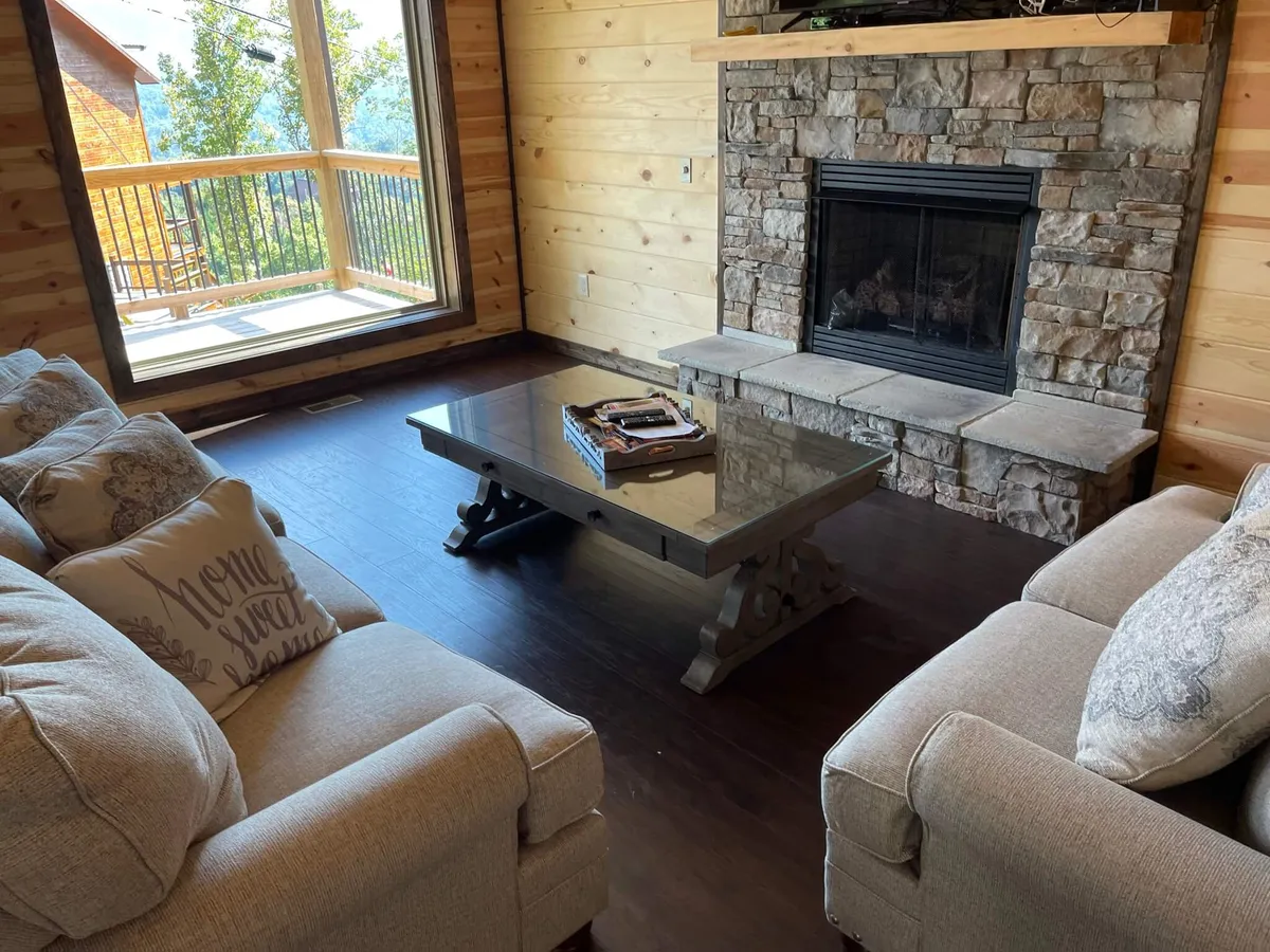 A bright, spacious living room featuring a prominent fireplace, a welcoming coffee table, and a large window that frames a scenic view. The space is arranged to invite group gatherings, relaxation, and conversation, with comfortable seating and natural light enhancing the inviting mood.
