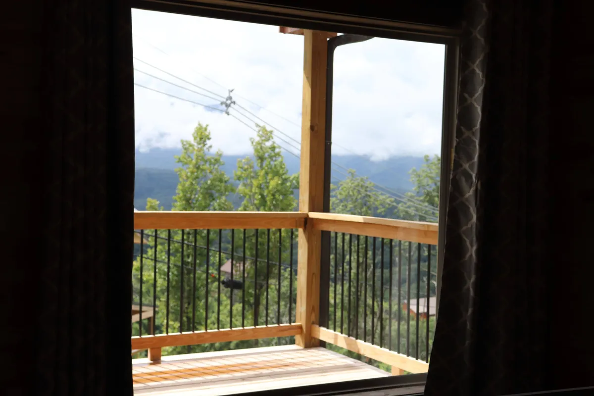 A wooden balcony with curtains frames a breathtaking view of lush trees and distant mountains. The space is inviting for morning coffee, reading, or simply soaking in the serene natural setting. The ambiance is relaxed, emphasizing the connection between the cabin and the outdoors.
